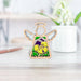 A small, Pansy Ornament 3.5" Angel Figurine from the Mother's Angels® collection by Forged Flare® stands on a light-colored wooden surface, showcasing intricately cut wings and a floral design painted in yellow, purple, and green. An out-of-focus white pot with green leaves and white flowers appears in the background.