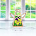 A 3.5-inch wooden angel figurine from the Mother's Angels® collection by Forged Flare®, known as the Pansy Ornament, is placed on a white surface. This intricate piece features delicate cut-outs and a floral design in green and purple at its center. The background offers a serene, blurred view of bright green scenery and large windows.