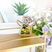 A wooden angel ornament from the Mother's Angels® collection by Forged Flare®, featuring wings, a halo, and a beautiful yellow and purple pansy flower design, is elegantly displayed on a gold-tiered shelf. In the background, potted plants and white flowers add to the decorative ambiance. This 3.5" Pansy Ornament makes for thoughtful flower gifts suitable for any occasion. The setting appears bright and decorative.