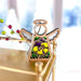 Forged Flare® presents the Pansy Ornament | 3.5" Angel Figurine from the Mother's Angels® collection, a charming wooden Christmas decoration. This angel-shaped ornament features a hollow center showcasing a delicate yellow and purple pansy flower and is equipped with a braided linen string for hanging. Positioned on a gold-colored surface with blurred festive decorations in the background, this exquisite piece makes an ideal addition to your floral decor.