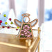 The Pink Evening Primrose Ornament, a 3.5" angel figurine from Mother's Angels® by Forged Flare®, features a wooden design with a hollow center adorned with pink evening primrose flowers. It stands gracefully on a gold surface and includes a braided linen string for hanging. The background showcases blurred bokeh lights and festive decorations, enhancing the holiday atmosphere.