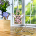 A Pink Evening Primrose Ornament from Forged Flare®'s Mother's Angels® collection, featuring a 3.5" wooden angel figurine with intricate cut-out designs and pink evening primrose flower accents, hangs elegantly on a stand. Set against a background showcasing a window with lush green outdoor scenery, a wooden pencil holder, and a vase of purple flowers on a wooden surface, this charming ornament makes for an ideal last-minute Mother's Day gift for mom.