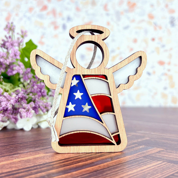 The American Flag Ornament by Forged Flare®, measuring 3.5 inches, is a charming wooden angel figurine from the Mother's Angels® collection. It features delicate wings and a halo, with a beautiful design of the American flag adorning its body. The scene is set against a backdrop of purple and white flowers arranged on a wooden surface against a light, speckled wall. This ornament makes an ideal patriotic gift for the holiday season.