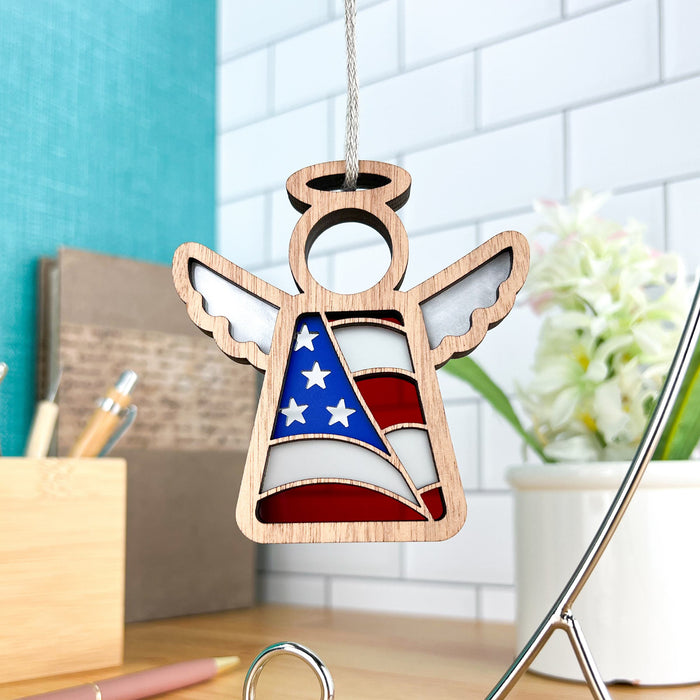 The American Flag Ornament from Forged Flare® features a 3.5" wooden angel figurine from the Mother's Angels® collection, with wings outstretched and a stained glass–style design of the American flag on its body. This patriotic gift is perfect as a Christmas tree ornament. In the background, you can see a desk with office supplies and a potted plant.