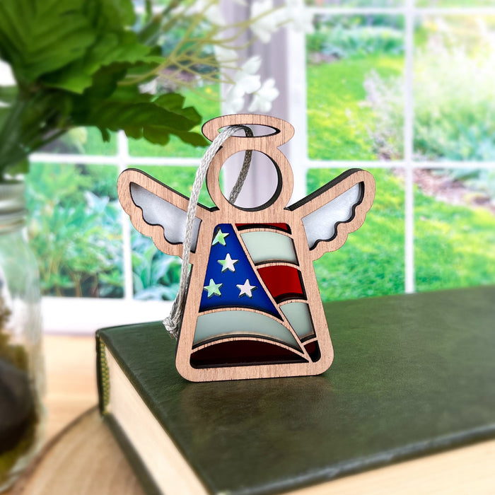 An American Flag Ornament, specifically the 3.5" Angel Figurine from the Mother's Angels® collection by Forged Flare®, is placed on a green book. The ornament features wooden construction and stained glass–style pieces in the colors of the American flag. In the background, a sunlit window reveals greenery outside, complemented by a nearby vase filled with green leaves.