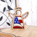 An American Flag Ornament, specifically the 3.5" Angel Figurine from the Mother's Angels® collection by Forged Flare®, is placed on a green book. The ornament features wooden construction and stained glass–style pieces in the colors of the American flag. In the background, a sunlit window reveals greenery outside, complemented by a nearby vase filled with green leaves.