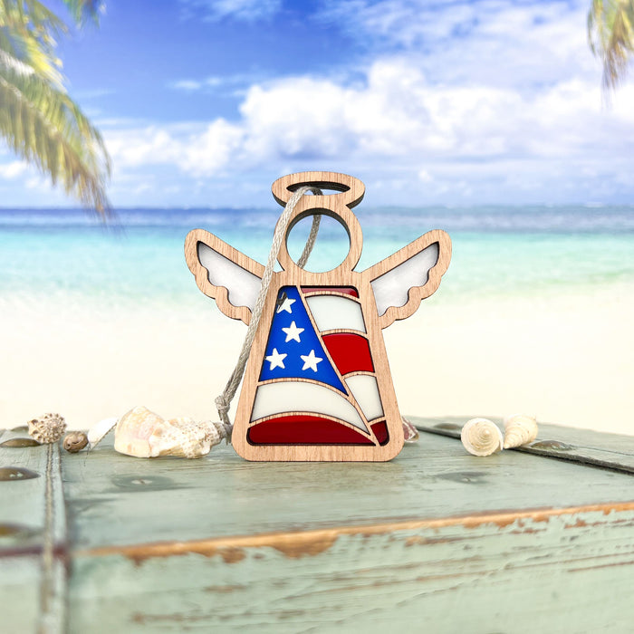 Displayed on a rustic surface adorned with seashells, the American Flag Ornament Angel Figurine from Mother's Angels® by Forged Flare® combines a small wooden angel with an intricate stained glass–style design of the American flag, showcasing stars and stripes. Ideal as a patriotic gift, it is set against a serene beach scene with turquoise water and palm trees in the background.