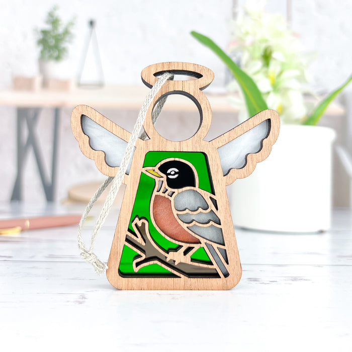 The American Robin Ornament from Forged Flare's Mother's Angels® collection features a vibrant American robin design at the center of a wooden, angel-shaped figurine with white wings and a green background. Measuring 3.5 inches and equipped with a loop for hanging, this charming ornament is photographed on a white surface with blurred background elements like a plant and pens, making it an ideal gift for bird lovers.