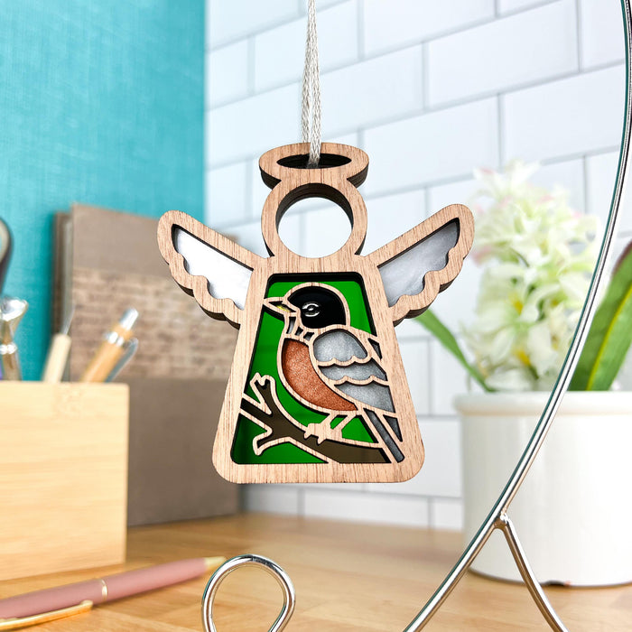 The American Robin Ornament from Forged Flare® is a stunning 3.5-inch angel figurine from the Mother's Angels® collection. Shaped like an angel with intricate cutouts, it features an American robin in black, white, and red perched on a branch against a green background. The ornament hangs by a string, with a blurred desk and various stationery items in the backdrop—making it the perfect gift for bird lovers.