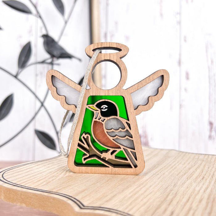 An American Robin Ornament from the Forged Flare® Mother's Angels® collection, featuring a 3.5" angel figurine with a detailed image of a bird perched on a branch, rests on a wooden surface. The green background and cut-out halo at the top add to its charm. In the blurred background is another bird ornament, making it an ideal gift for bird lovers.