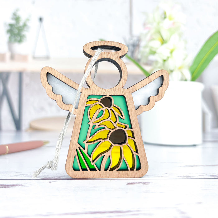 The Black-Eyed Susan Ornament, a 3.5" angel figurine from Forged Flare's Mother's Angels® collection, rests on a white surface. This wooden decoration, ideal as a Christmas tree ornament, showcases a stained glass–style design with two yellow flowers featuring dark brown centers and green leaves. In the background, blurred objects such as a plant, pen, and cup contribute to the cozy scene.