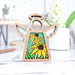 The Black-Eyed Susan Ornament, a 3.5" angel figurine from Forged Flare's Mother's Angels® collection, rests on a white surface. This wooden decoration, ideal as a Christmas tree ornament, showcases a stained glass–style design with two yellow flowers featuring dark brown centers and green leaves. In the background, blurred objects such as a plant, pen, and cup contribute to the cozy scene.