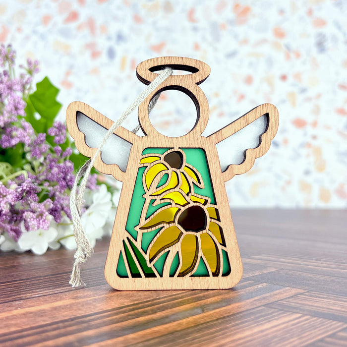 A 3.5" angel figurine from Mother's Angels® by Forged Flare®, adorned with intricate cutouts and Black-Eyed Susan accents, is displayed on a polished wooden surface. The delicately crafted wings and halo feature fine details, and a braided linen string is attached for hanging. In the background, there is a soft-focus floral arrangement against a blurred multicolored backdrop.