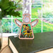 A Forged Flare® Mother's Angels® 3.5" Angel Figurine, featuring a wooden Black-Eyed Susan ornament with intricate cutouts of a yellow flower and green leaves, sits atop a closed green book. The Christmas tree ornament has a string for hanging. Behind it, a large window reveals a lush outdoor garden scene. A vase with flowers is positioned on the left side.