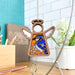 The Blue Jay Ornament from the Mother's Angels® collection by Forged Flare® is a 3.5" angel figurine featuring a stunning bluebird and flower design with stained glass–style details. Perfect as a gift for bird lovers, this wooden ornament hangs gracefully from a string in well-lit spaces like kitchens or workspaces adorned with white subway tiles, plants, and various office supplies on wooden surfaces.