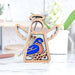 Introducing the Bluebird Ornament from Forged Flare®, a 3.5" angel figurine from the Mother's Angels® collection. This wooden angel ornament features gracefully detailed wings and a vibrant inlaid design of a bluebird perched on a branch with red berries. A looped string is attached at the top, making it easy to hang this perfect gift for bird lovers. It is beautifully displayed on a light-colored table with plants and decor in the background.