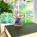 The Bluebird Ornament, a 3.5" Angel Figurine from Forged Flare's Mother's Angels® collection, is displayed on a closed book and features intricate stained glass detailing of a bluebird. A lush garden is visible through the large window in the background, making it an ideal gift for bird lovers. A vase with white flowers can be partially seen on the left side of the scene.