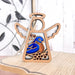 A Forged Flare® Mother's Angels® Bluebird Ornament, a 3.5" wooden angel figurine featuring wings and a halo, showcases a vibrant bluebird design on its body. It is set against a light wooden surface with a metal bird decoration in the blurred background, making it an ideal gift for bird lovers.