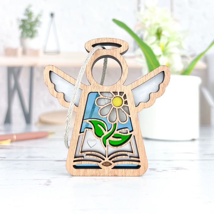 The Book Lovers Ornament by Forged Flare® is a 3.5" wooden angel figurine from the Mother's Angels® collection, featuring a cut-out design with stained glass–style detailing that depicts a daisy flower and leaves growing from an open book. Ideal as a Christmas tree ornament or a thoughtful gift for book club enthusiasts, the background captures blurred home decor items such as a potted plant and writing utensils.