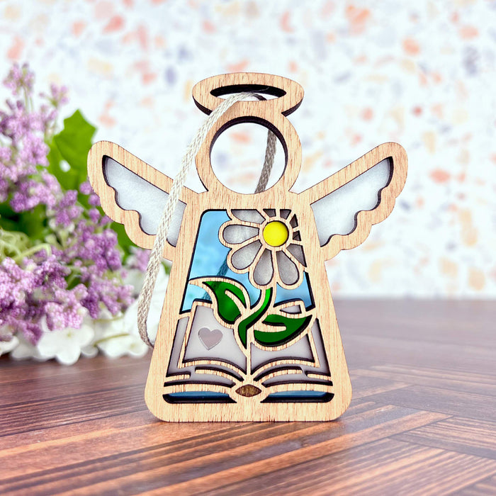 The Book Lovers Ornament from Forged Flare®, measuring 3.5 inches, showcases a wooden angel figurine with elegant wings and a halo. It features a vibrant stained glass design at its center, depicting a yellow and white flower surrounded by green leaves. The background highlights purple flowers against a wooden surface with floral wallpaper—making it the perfect gift for authors or book club members.