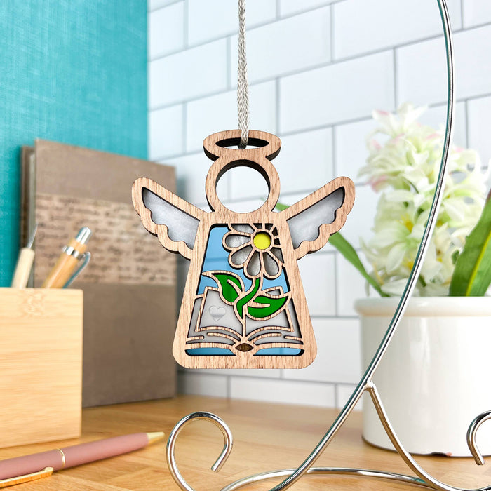 The Book Lovers Ornament by Forged Flare®, a 3.5" angel figurine from the Mother's Angels® collection, showcases wooden construction with wings and a vibrant inlay design featuring an open book and a daisy with green leaves. Perfect as a gift for book club members or authors, this ornament hangs on a string from a metal display stand placed on a wooden desk, set against the blurred background of a green wall, books, and a plant.