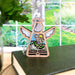 The Book Lovers Ornament by Forged Flare® showcases the 3.5" Angel Figurine from Mother's Angels®, featuring intricate cut-out details of a flower and leaves, elegantly positioned on a closed book. The background displays a bright window with a glimpse of a lush, partially blurred green garden. Perfect as a book club gift or Christmas tree ornament, the scene is complemented by part of a leafy plant in a glass container visible on the left side.