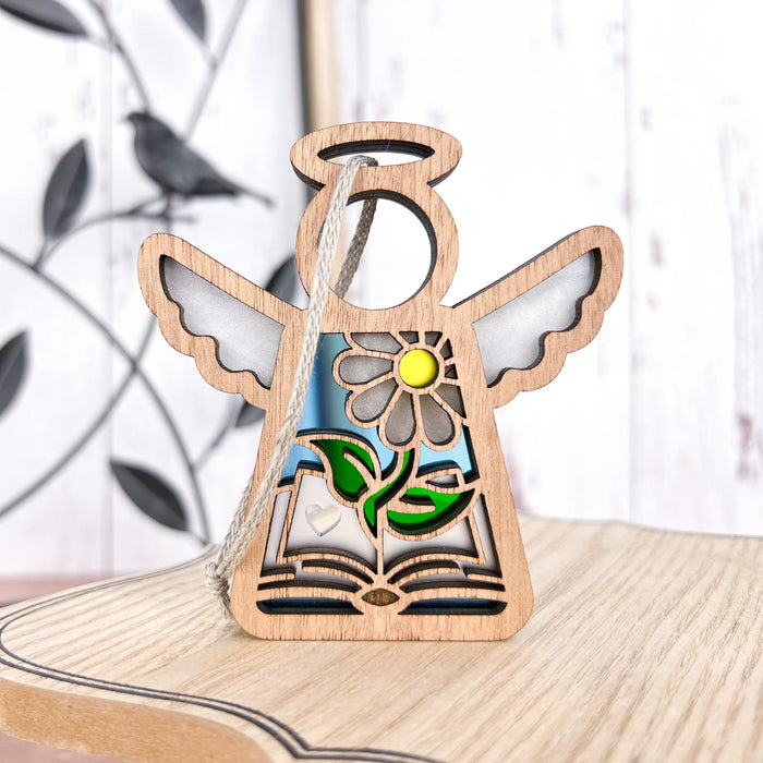 A wooden Book Lovers Ornament from the Mother's Angels® collection by Forged Flare® stands on a light wood surface. The 3.5" angel figurine, ideal as a Christmas tree ornament, features white wings and showcases a cutout design with a blooming flower and an open book painted in vibrant colors. A bird and branch silhouette decorates the background.