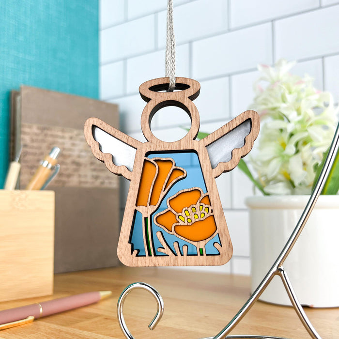 A California Poppy Ornament from the Mother's Angels® collection by Forged Flare® features an angel figurine with white wings, adorned with stained glass–style California poppy flowers in orange and yellow. Measuring 3.5 inches, it hangs from a light-colored string against a backdrop of a tiled wall, alongside a desk filled with books, pens, and a potted plant.