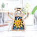 A small Celestial Sun Ornament from Forged Flare® stands on a white table. This 3.5" angel figurine from Mother's Angels® features a wooden design with a halo, outstretched wings, and a brightly colored sun with yellow and orange rays against a blue background. In the blurred background, there are plants and a white wall.