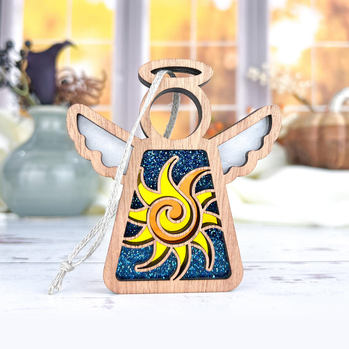 A Celestial Sun Ornament from Forged Flare® (part of the Mother's Angels® collection) sits on a table. The 3.5" angel figurine has a halo and outstretched wings, with its body showcasing a vibrant design: a yellow sun with swirling rays against a blue starry background. In the softly blurred backdrop, autumnal decorations and a window can be seen, making it an ideal unique Christmas ornament.