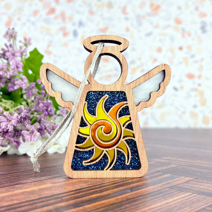 The Celestial Sun Ornament, a 3.5" angel figurine from Mother's Angels® by Forged Flare®, is a decorative wooden Christmas ornament with a halo and colorful sunburst design on its body. The angel hangs by a string and is displayed on a wooden surface, with a background adorned with purple flowers and green leaves.