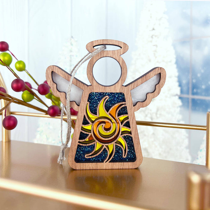 A charming Celestial Sun Ornament from Forged Flare's® Mother's Angels® collection, depicting a 3.5" wooden angel figurine with outspread wings and a vivid yellow and orange swirling sun design on its body, stands on a shelf. The background is adorned with holiday decor featuring red and green berries alongside snowy trees visible through a frosty window, making this delightful piece the perfect Christmas ornament.