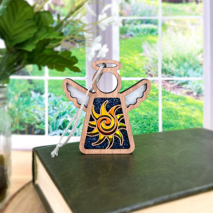 A decorative wooden 3.5" angel figurine from the Mother's Angels® collection by Forged Flare®, featuring a colorful celestial sun design on its body, sits atop a closed hardcover book. The background showcases a blurred view of a lush green garden through a large window, with a jar containing green leaves partially visible on the left.