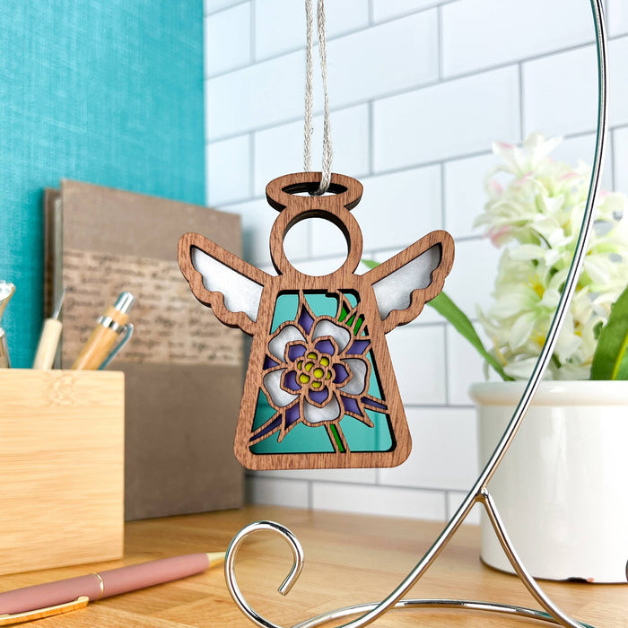 The Columbine Ornament, a 3.5" angel figurine from the Mother's Angels® collection by Forged Flare®, hangs gracefully from a braided linen string. Featuring delicate white wings and a charming floral pattern at its center, it is displayed in an organized and bright workspace with a notepad, pens, and a small white vase filled with flowers in the background. This setting perfectly showcases the enchanting Christmas tree ornament.