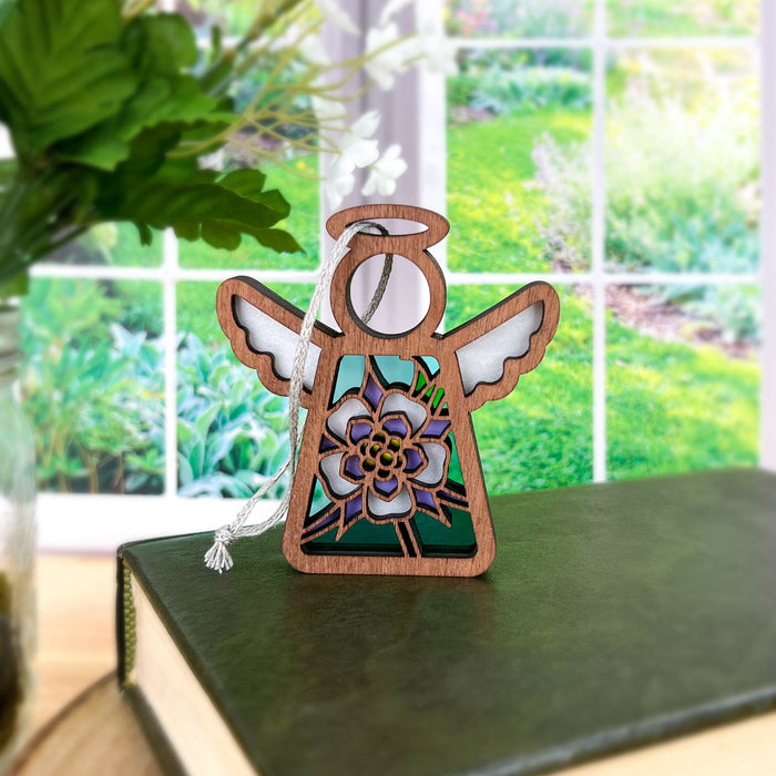 A small Columbine Ornament, a 3.5" wooden angel figurine from the Mother's Angels® collection by Forged Flare®, with intricate wings and a flower design in the center, stands on a closed book. In the background, a large window reveals a lush green garden scene with various plants. A vase with greenery is partially visible on the left, enhancing the serene ambiance of this flower decor.