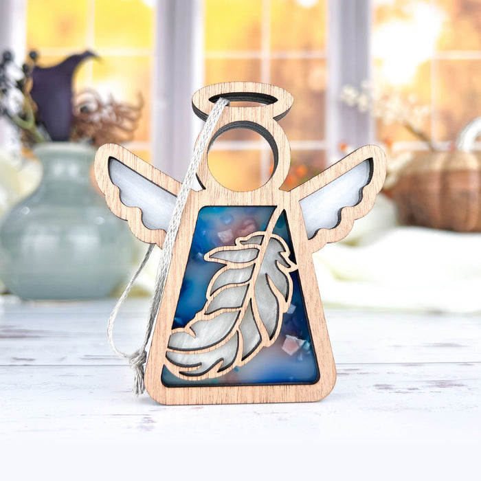 The Feather Ornament | 3.5" Angel Figurine from the Mother's Angels® collection by Forged Flare® is a heartfelt memorial gift featuring a wooden angel with white wings and a hanging string. The cut-out design showcases an inset of blue, marbled pattern and a feather in the center. The softly lit background includes vases and blurred window panes, adding to its touching appeal.