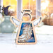 The Feather Ornament | 3.5" Angel Figurine from the Mother's Angels® collection by Forged Flare® is a heartfelt memorial gift featuring a wooden angel with white wings and a hanging string. The cut-out design showcases an inset of blue, marbled pattern and a feather in the center. The softly lit background includes vases and blurred window panes, adding to its touching appeal.