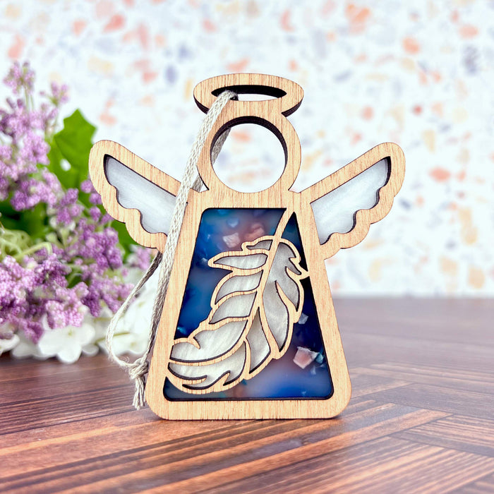 The Feather Ornament | 3.5" Angel Figurine from Mother's Angels® by Forged Flare® features a wooden angel with a halo, wings, and a translucent blue body adorned with a feather design. It rests on a wooden surface with a small bouquet of purple flowers and greenery in the background, making it an ideal mindfulness gift for any occasion.