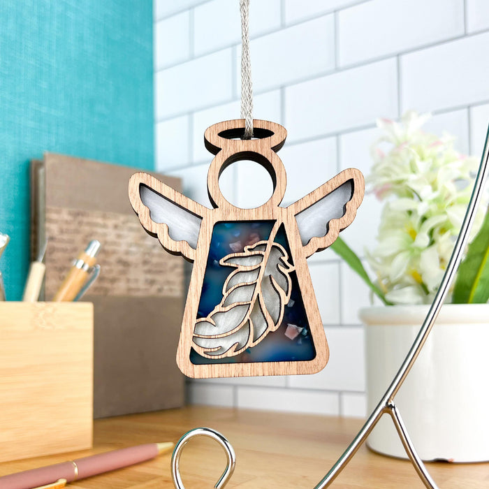 The Feather Ornament from Forged Flare® is a 3.5" wooden angel figurine featuring a marbled blue section and a delicate feather design, hanging elegantly on a string. In the background, there's a desk adorned with a penholder, some books, lush greenery, and a pristine white-tiled wall. This charming piece from the Mother's Angels® collection makes for an ideal mindfulness gift or Christmas tree ornament.