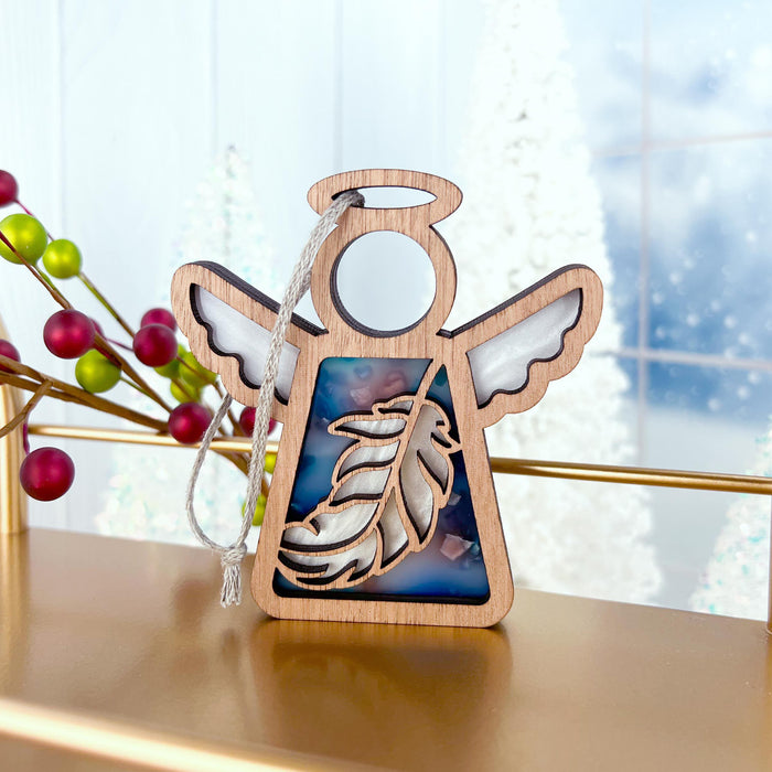 The Feather Ornament | 3.5" Angel Figurine from Mother's Angels® by Forged Flare® features a wooden angel ornament with white wings, set against a blue multicolored background adorned with a feather design in the middle. Perfect as a Christmas tree ornament or mindfulness gift, it hangs beautifully against a snowy window decorated with festive red berries and green leaves.