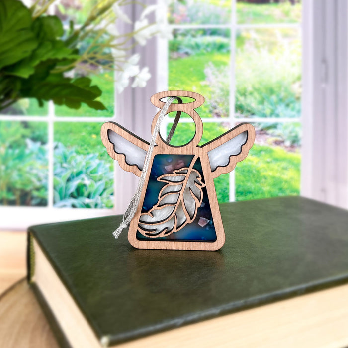 An intricate 3.5" angel figurine from Forged Flare®'s Mother's Angels® collection, the Feather Ornament, depicts a wooden angel with a halo and delicate feather detailing on its wings and body, standing gracefully on a closed book. Ideal as a mindfulness gift, it is set against the backdrop of a bright green garden seen through an expansive window.