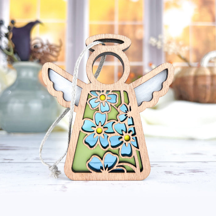 The Forget-Me-Not Ornament, a 3.5" angel figurine from the Mother's Angels® collection by Forged Flare®, is displayed on a tabletop. The wooden angel features a hollow circular head, white wings, and a floral dress design adorned with blue forget-me-nots against a green background. The scene includes vases, baskets, dried flowers, and is lit by sunlight streaming through the window.