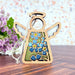 The Forget-Me-Not Ornament, a 3.5" angel figurine from the Mother's Angels® collection by Forged Flare®, features intricate blue forget-me-not floral designs and a braided linen string for hanging. It is placed on a wooden surface with a decorative arrangement of purple blooms in the background and a speckled pattern on the wall.