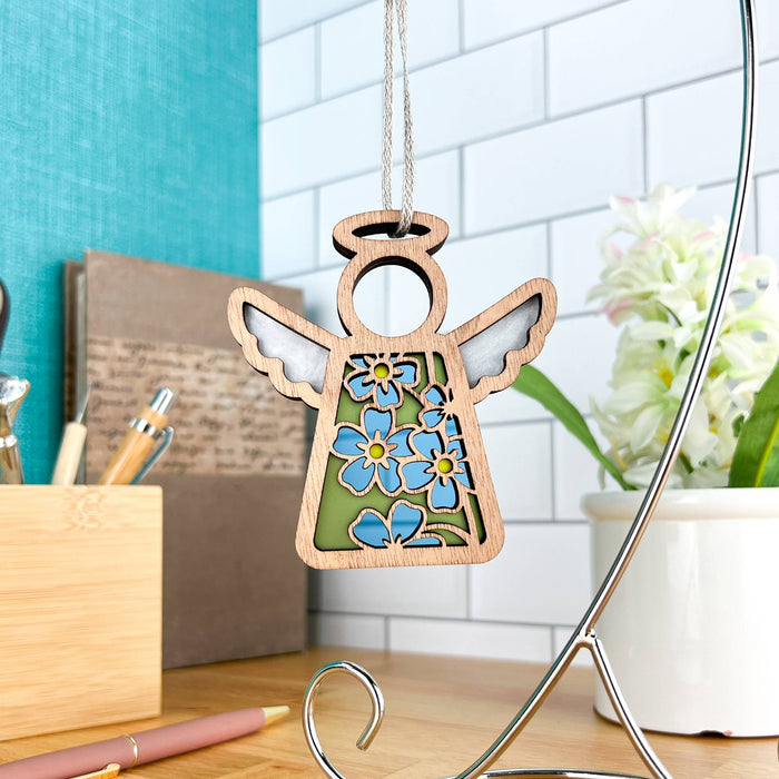 The Forget-Me-Not Ornament | 3.5" Angel Figurine from the Mother's Angels® collection by Forged Flare® features a wooden angel adorned with blue forget-me-not flower decorations and white wings, hanging on a stand in a home office setting. The background showcases a tiled wall, desk accessories, a beige pen, a note holder, and a potted plant.