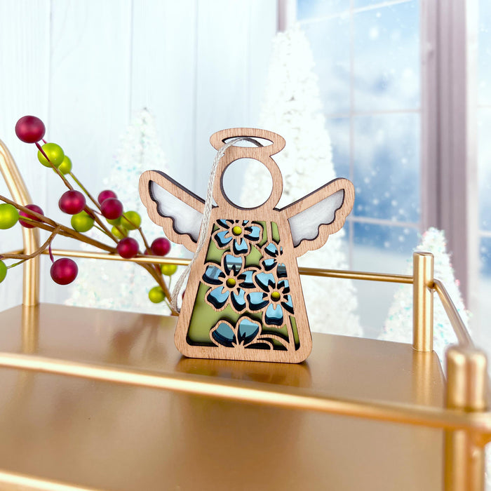 A Forget-Me-Not Ornament from the Mother's Angels® collection by Forged Flare®, featuring a 3.5" wooden angel figurine with floral designs, stands on a golden cart decorated with forget-me-nots. In the background, branches with red berries and green leaves are visible against a snowy scene through a window.