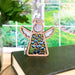 The Forget-Me-Not Ornament by Forged Flare®, a 3.5" angel figurine from the Mother's Angels® collection, features white wings and floral cut-out patterns. It stands on a closed book against a bright window. The green foliage visible through the window creates a natural, peaceful background that beautifully complements the delicate forget-me-not flowers.