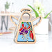 A Forged Flare® Mother's Angels® Fuchsia Ornament, a 3.5" wooden angel figurine adorned with a vibrant stained glass–style purple and red fuchsia, stands on a white tabletop. The angel features a braided linen string for hanging, white wings, and a halo. Blurred plant and desk items can be seen in the background.