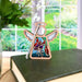 The Fuchsia Ornament, a 3.5" angel figurine from Mother's Angels® by Forged Flare®, showcases colorful stained glass–style wings and body, perched atop an old closed book. Behind this charming ornament is a bright window offering a view of a lush green outdoor garden.