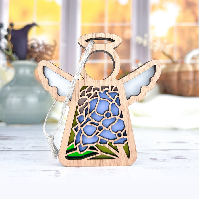 The Hydrangea Ornament, a 3.5" angel figurine from the Mother's Angels® collection by Forged Flare®, is displayed on a white surface. This wooden decoration features a stained glass–style design with blue flowers, along with white wings and a halo. The background showcases a blurred, warm-toned indoor scene with windows, bottles, and vases.