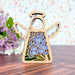 A stunning 3.5" angel figurine from the Mother's Angels® collection by Forged Flare® stands gracefully on a wooden surface. This Hydrangea Ornament features intricate wood carvings and stained glass–style floral wings, adorned with a braided linen string for hanging. In the background, a bouquet of purple flowers against a light-colored speckled wall creates an enchanting setting, making it an ideal addition to any Christmas tree or angel figurine collection.